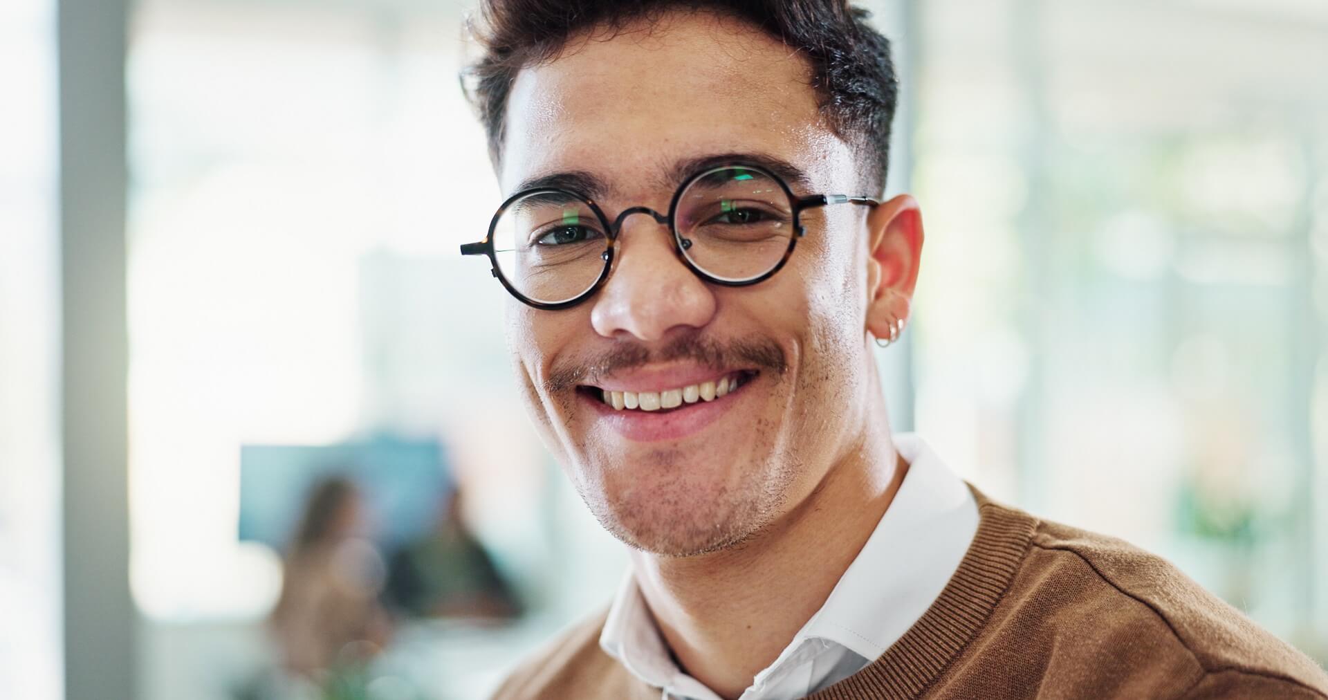 Mann mit Brille am Schreibtisch vor unscharfem Bildschirm im Hintergrund